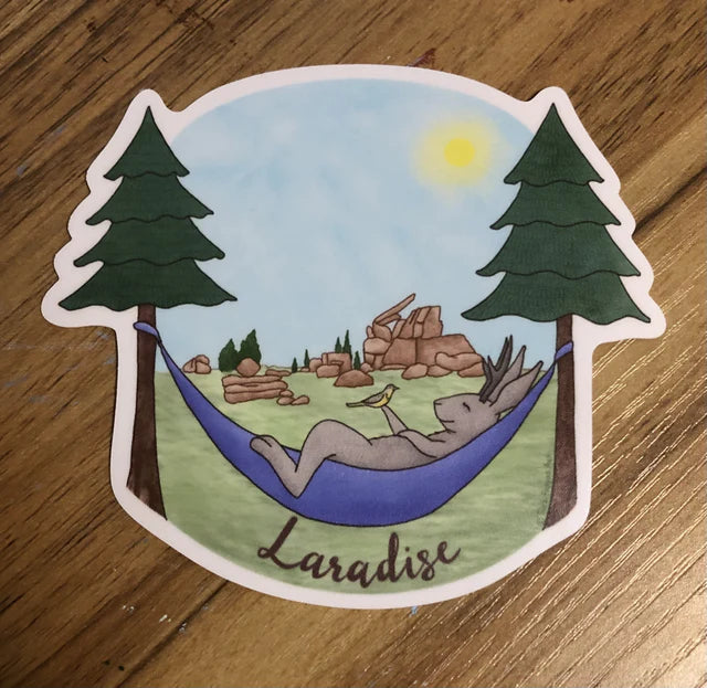 cute whimsical sticker of a Jackalope laying in a hammock holding a meadowlark bird.  Rock formations from Vedauwoo Wyoming are in the background. Laradise is written at the bottom