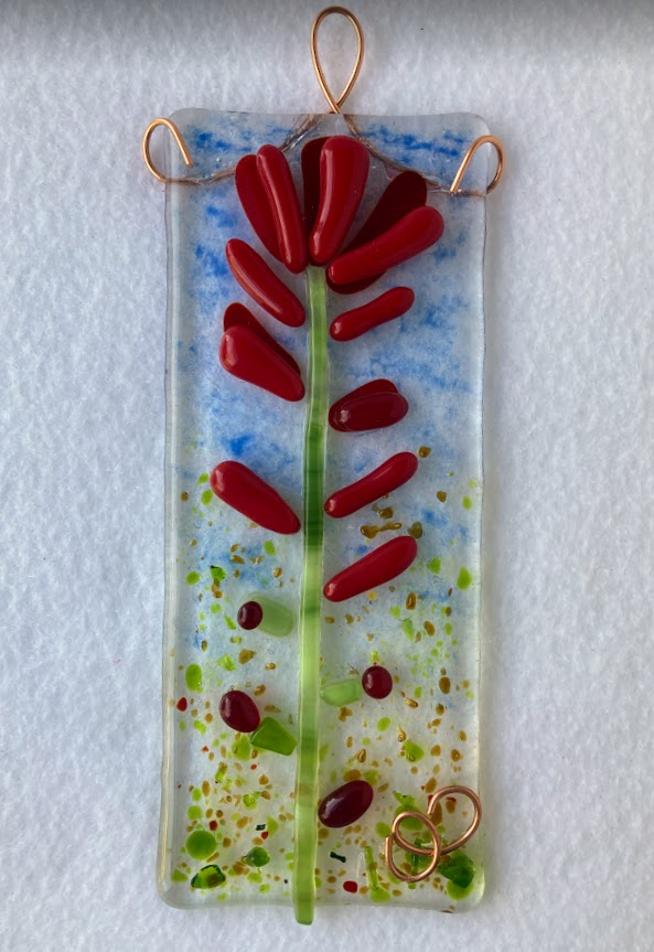 fused glass suncatcher. red glass creates the petals of the Indian paintbrush flower. green glass creates the stem.  blue and green in the glass make a sky above and grass below. copper wire embelishments also give the place for a hanger.